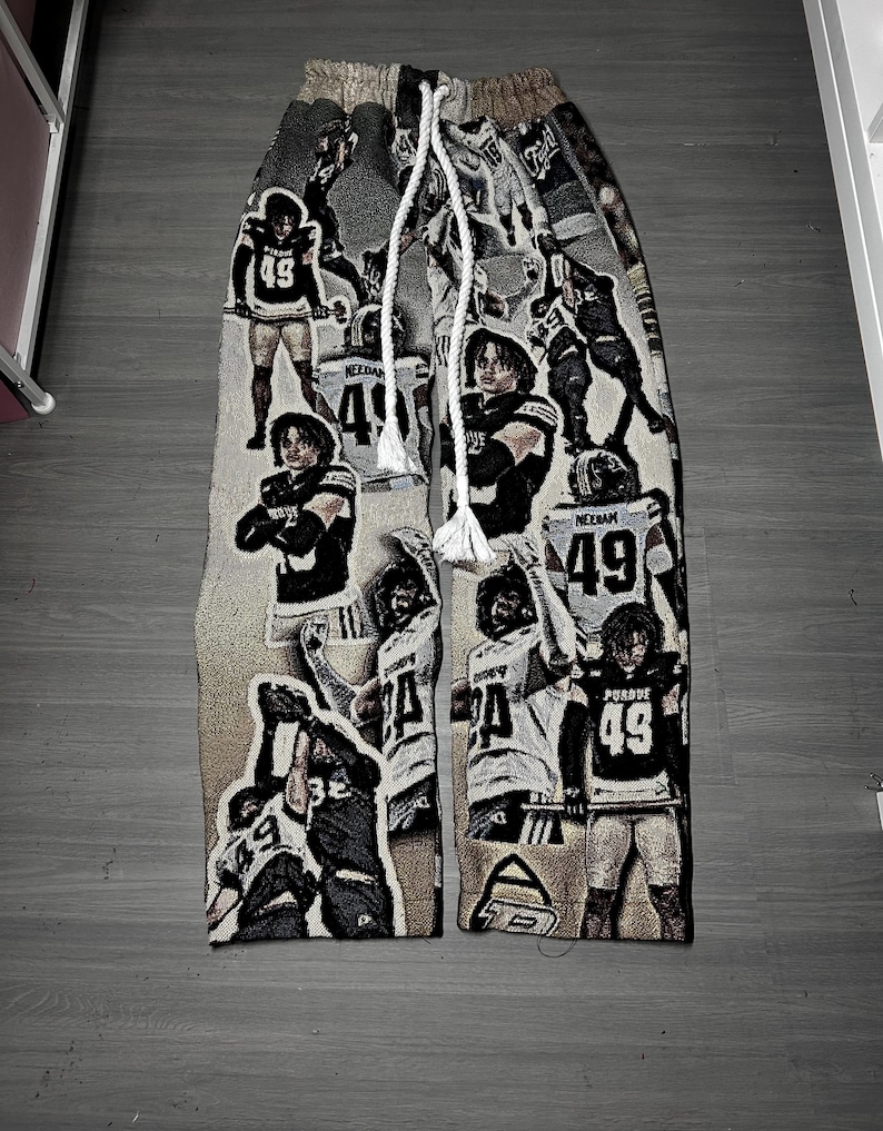 May include: Tapestry-style sweatpants featuring a repeating pattern of football players in action. The pants are primarily beige and brown, with a white drawstring. The players are wearing black and white uniforms with the number 49.