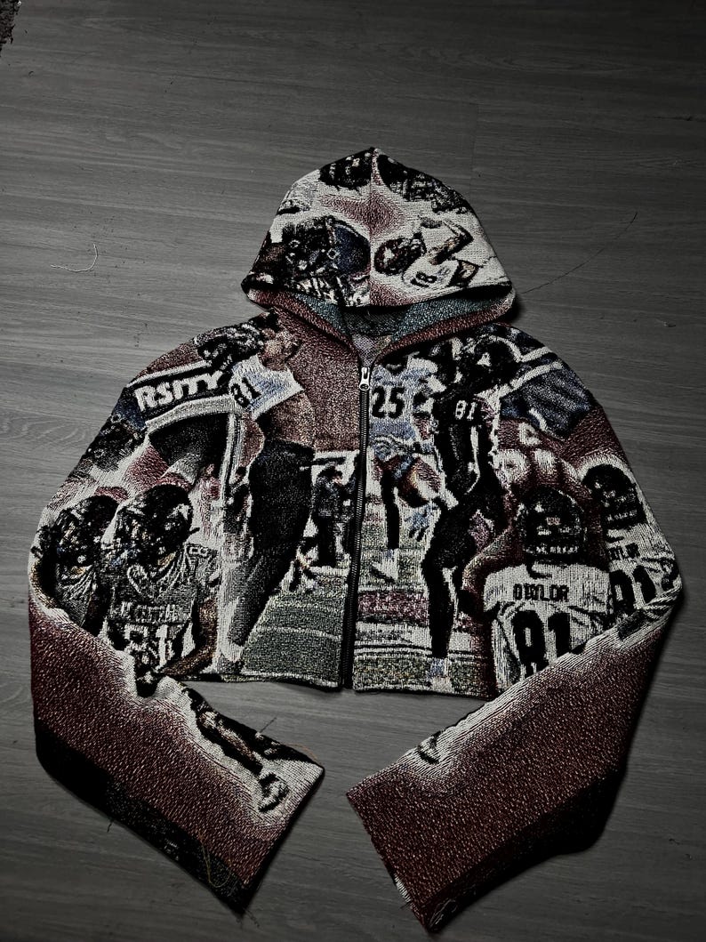 May include: Cropped zip-up hoodie with a tapestry design depicting football players. The jacket, with a hood, is predominantly black, white, and burgundy. The word "RSITY" is visible on the front.