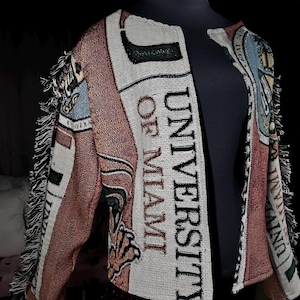May include: A woven jacket featuring the text "UNIVERSITY OF MIAMI" in black. The jacket is predominantly white, orange, and brown, with fringed edges. It is displayed on a black mannequin, with the text clearly visible.