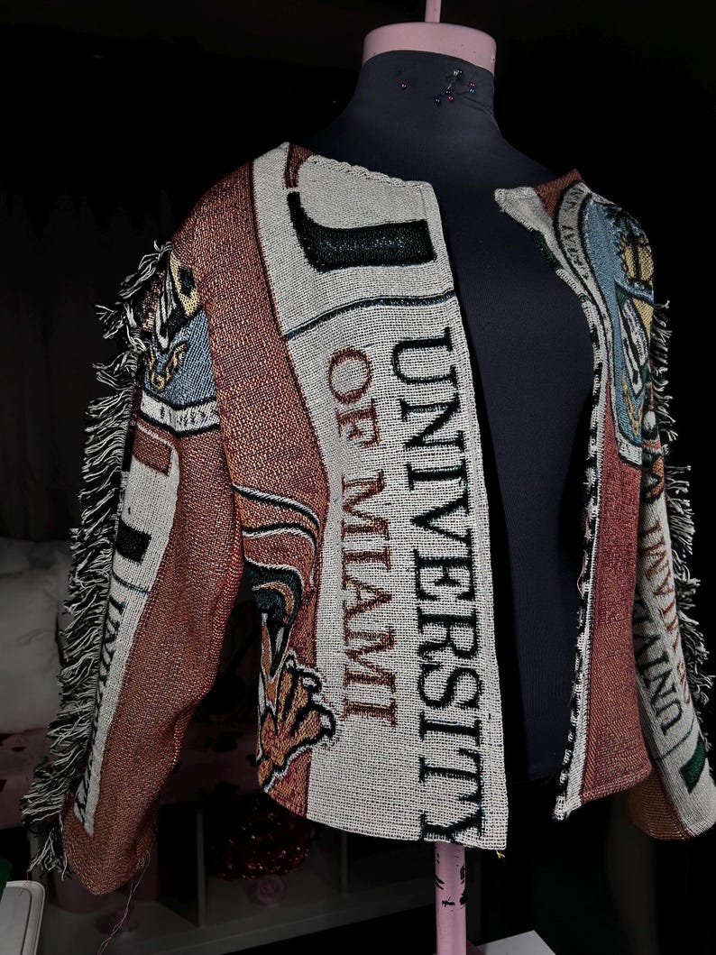 May include: A woven jacket featuring the text "UNIVERSITY OF MIAMI" in black. The jacket is predominantly white, orange, and brown, with fringed edges. It is displayed on a black mannequin, with the text clearly visible.