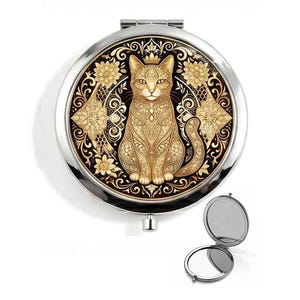 May include: A round silver compact mirror featuring a gold-toned cat illustration with intricate filigree patterns. The cat is centered within a decorative border of floral motifs and geometric shapes. A smaller open version of the mirror is shown below.