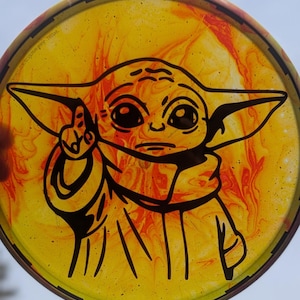 Puede incluir: Un disco redondo amarillo y naranja con una ilustración negra de un personaje con ojos grandes y orejas puntiagudas. El personaje lleva una túnica y tiene un dedo levantado. El disco tiene un borde oscuro.