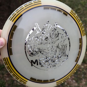 May include: A white disc golf disc with a yellow and black rim. The disc features a silver metallic design of a robot with the text "MECHA" and "CENTRAL COAST DISC GOLF" on it.