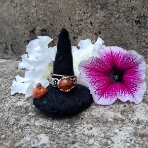 May include: A silver ring featuring a round, polished amber stone set in a decorative metal band, positioned on a black felt cone. The ring is accompanied by a small orange felt pumpkin and surrounded by white and vibrant magenta petunia flowers.
