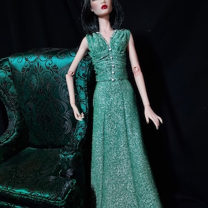 May include: A fashion doll wearing a shimmering green gown with a V-neckline and silver button details. The doll has black hair and red lipstick. A green patterned chair is to the left of the doll. The background is black.