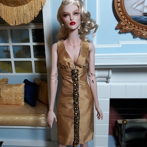 May include: A fashion doll wearing a gold dress with a beaded embellishment down the front. The doll has blonde hair and blue eyes.