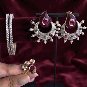 May include: A jewellery set featuring two silver-toned bangles encrusted with clear stones, a pair of crescent-shaped earrings with deep red teardrop accents, small green and red stones, and white beads, and a gold-toned ring with three round stones.