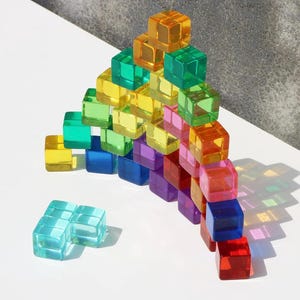 May include: A colorful arrangement of translucent cubes in various colors, including yellow, green, blue, purple, and red. The cubes are stacked to form a pyramid shape, with a few cubes separated from the main structure. The cubes cast shadows on a white surface.