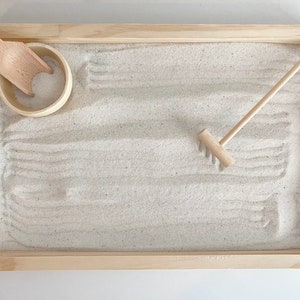 May include: A wooden tray with a white sand surface, a small wooden scoop, and a wooden rake. The tray is perfect for creating calming sand art.