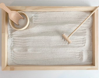 May include: A wooden tray with a white sand surface, a small wooden scoop, and a wooden rake. The tray is ideal for creating calming sand art.