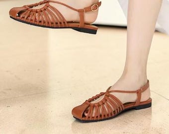 May include: A pair of tan brown flat sandals featuring a woven cage-style upper with thin straps, a rounded toe, and an adjustable ankle strap with a small gold-toned metal buckle closure, worn with a flowing white skirt.