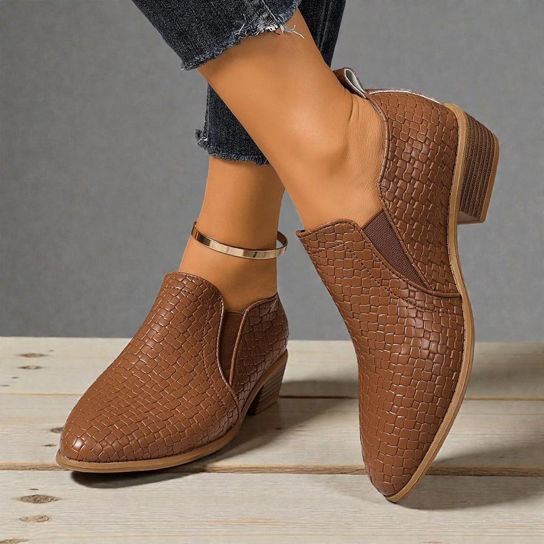 Pu&ograve; includere: Stivaletti marroni con una trama intrecciata e un tacco a blocco basso. Gli stivaletti hanno la punta affusolata e pannelli laterali elastici. Le scarpe sono mostrate su una superficie di legno.