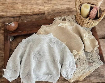 May include: Two long-sleeved knit jumpers, one in light grey and one in beige, each featuring a crew neckline and multiple sheer organza ribbon bows attached to the front and sleeves. The jumpers are displayed on a wooden surface.