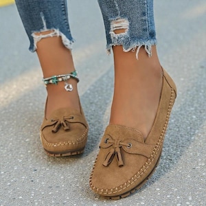 May include: Close-up of brown suede loafers with a bow detail and stitched trim. The shoes are worn with distressed blue jeans. The loafers have a rounded toe and a rubber sole.
