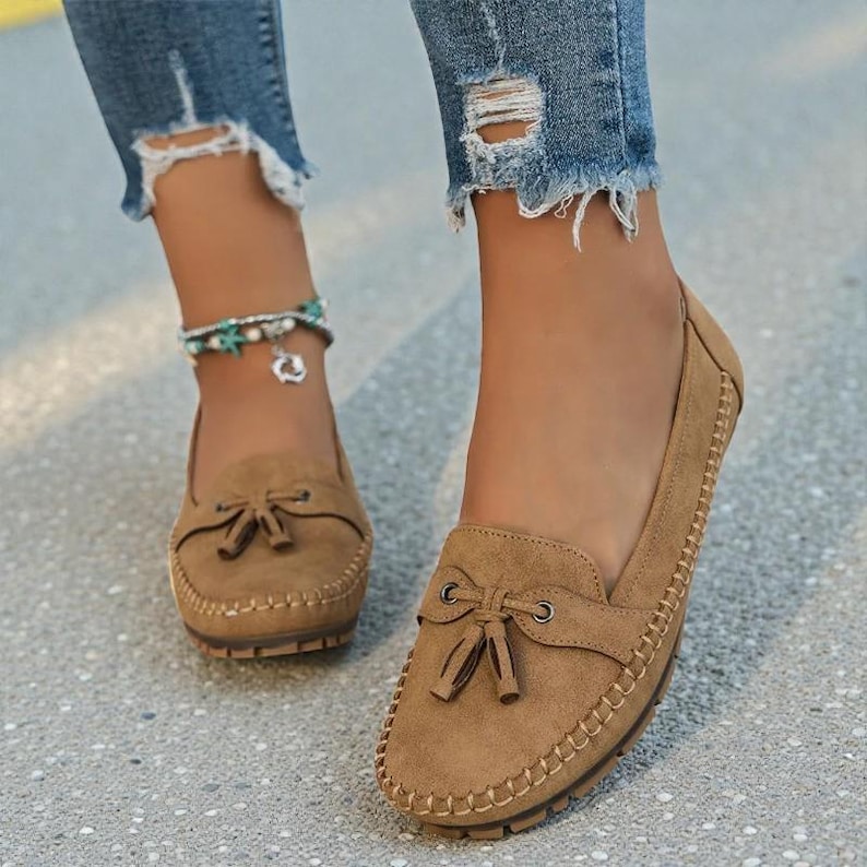May include: Close-up of brown suede loafers with a bow detail and stitched trim. The shoes are worn with distressed blue jeans. The loafers have a rounded toe and a rubber sole.