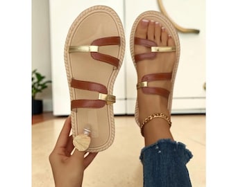May include: Flat sandals featuring a beige textured footbed with a braided edge. The upper consists of brown leather-like straps accented with metallic gold bars. One sandal is held in hand, while the other is worn on a foot.