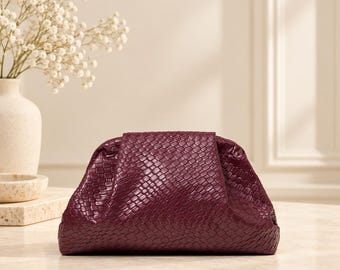 May include: A deep burgundy clutch bag featuring a woven, textured surface with a random pattern. The bag has a structured, rounded shape with a frame closure, displaying a consistent dark red colour throughout its exterior surface.