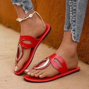 May include: Red leather sandals with a gold-colored accent. The sandals have a flat sole and a thong-style design. The sandals are worn with distressed denim jeans and a shell anklet.