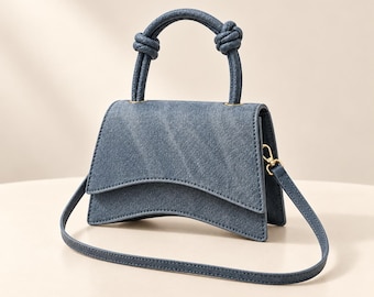 May include: A blue denim handbag featuring a curved bottom edge and a structured flap closure. The bag includes a top handle with knotted detailing on both sides and a detachable, adjustable shoulder strap with gold-toned metal hardware.
