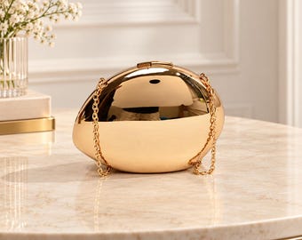 May include: A polished gold-tone clutch bag with an oval, egg-like shape and a smooth, reflective surface. The accessory features a thin, delicate gold-tone metal chain strap attached to the top, providing a sleek and minimalist appearance.