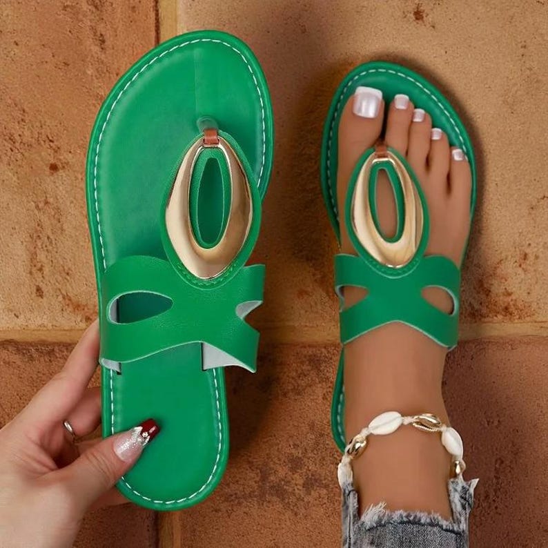 May include: Green sandals with a gold oval accent. The sandals have a thong-style design and a cut-out detail on the straps. The soles are green with white stitching. The sandals are shown on a person's foot.