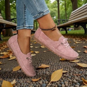 Puede incluir: Mocasines rosas de ante con dise&ntilde;o de cordones y costuras. Los zapatos tienen una puntera redondeada y suela de goma. La imagen muestra los zapatos puestos al aire libre sobre un camino de guijarros con hojas ca&iacute;das.