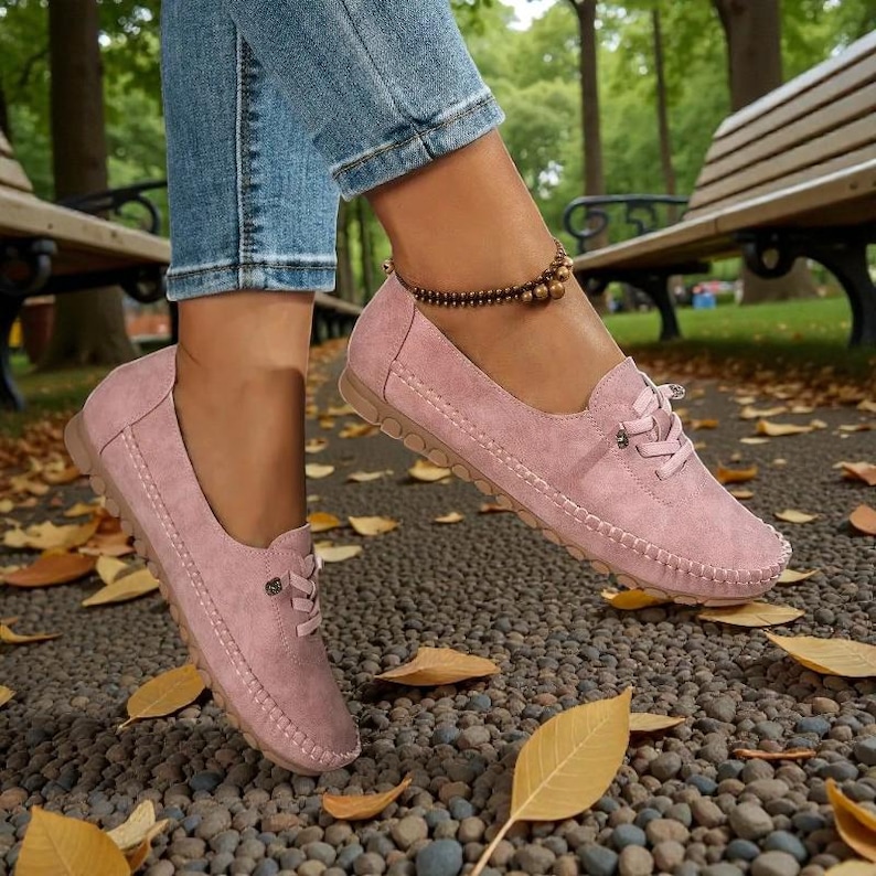Puede incluir: Mocasines rosas de ante con dise&ntilde;o de cordones y costuras. Los zapatos tienen una puntera redondeada y suela de goma. La imagen muestra los zapatos puestos al aire libre sobre un camino de guijarros con hojas ca&iacute;das.