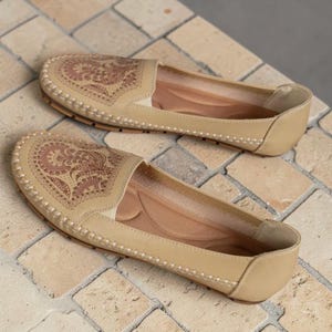 Op de afbeelding: Een paar beige instappers met een decoratief bloemmotief op de bovenkant. De schoenen hebben witte stiksels langs de randen en een neutraal gekleurde binnenkant. De schoenen zijn gemaakt van een zacht materiaal.