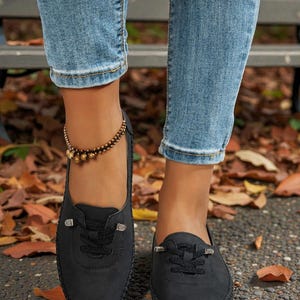 Puede incluir: Mocasines negros de ante sint&eacute;tico con costuras decorativas y cordones. Los zapatos tienen una punta redonda y una peque&ntilde;a hebilla decorativa en los cordones. Los zapatos se usan con vaqueros azules y una pulsera de tobillo con cuentas.