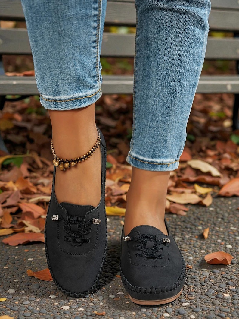 Puede incluir: Mocasines negros de ante sint&eacute;tico con costuras decorativas y cordones. Los zapatos tienen una punta redonda y una peque&ntilde;a hebilla decorativa en los cordones. Los zapatos se usan con vaqueros azules y una pulsera de tobillo con cuentas.