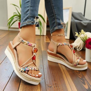 May include: Beige platform sandals with colorful braided straps and decorative beads. The sandals have a white sole and a woven jute-style trim. The straps feature floral patterns and the sandals are worn with blue jeans.