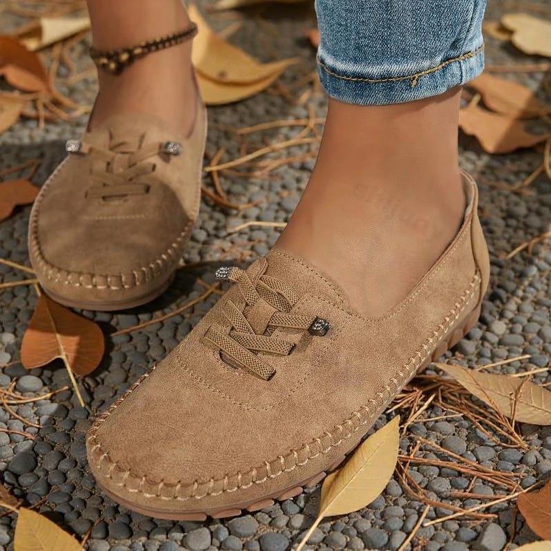 Puede incluir: Primer plano de mocasines de ante marr&oacute;n con costuras decorativas y detalles de cordones. Los zapatos tienen una puntera redondeada y una suela de goma. La imagen muestra los zapatos sobre una superficie de guijarros con hojas ca&iacute;das.