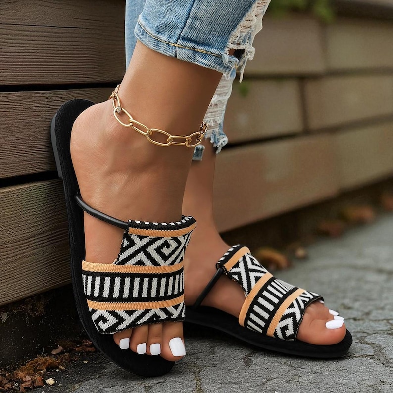 May include: Black and white patterned sandals with a black sole and a strap. The sandals have a geometric design in black, white, and tan. A gold chain anklet is also visible.