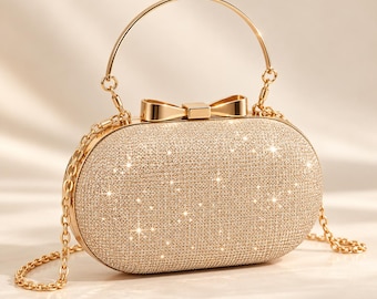 May include: An oval-shaped clutch bag covered in fine gold glitter, featuring a rigid gold-tone metal frame, a central decorative bow clasp, a thin circular top handle, and a detachable gold-tone chain shoulder strap.