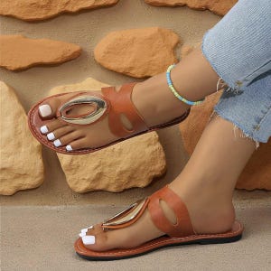 May include: Brown leather sandals with a gold-colored oval accent. The sandals have a thong-style design and are worn on a foot with white nail polish. The sandals are on a light brown surface.