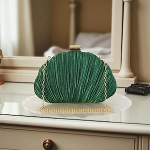May include: A dark green pleated clutch bag with a gold-coloured clasp and chain strap. The evening bag is displayed on a white plate. The clutch has a fan-like design, and is approximately 25 cm wide.