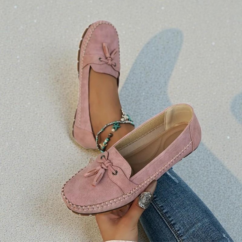 May include: Pink suede loafers with a tassel detail. The shoes have a stitched sole and a rounded toe. One shoe is on a foot with an ankle bracelet, and the other is held by a hand.
