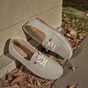 Puede incluir: Un par de mocasines de ante gris claro con cordones y costuras blancas. Los zapatos tienen una puntera redondeada y una suela marr&oacute;n. Los mocasines descansan sobre un lecho de hojas y hierba secas.