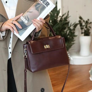 May include: A brown leather handbag with a gold clasp and a long strap. The bag has a top handle and a flap closure.