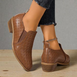 Pu&ograve; includere: Stivaletti marroni con un motivo strutturato a forma di rettile. Gli stivaletti hanno un tacco basso e un design slip-on con pannelli laterali elastici. &Egrave; visibile una cavigliera a catena dorata.