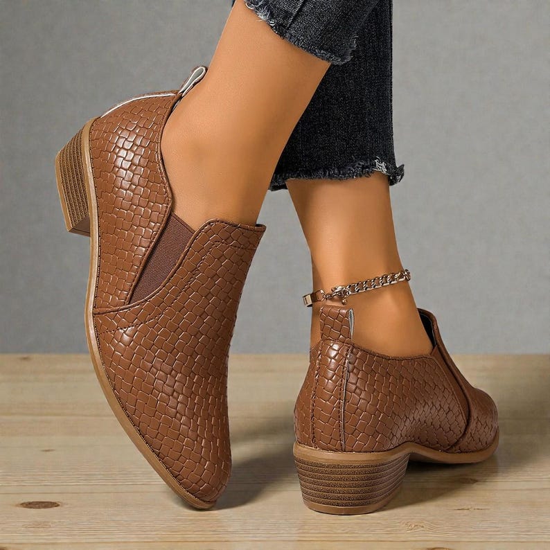Pu&ograve; includere: Stivaletti marroni con un motivo strutturato a forma di rettile. Gli stivaletti hanno un tacco basso e un design slip-on con pannelli laterali elastici. &Egrave; visibile una cavigliera a catena dorata.