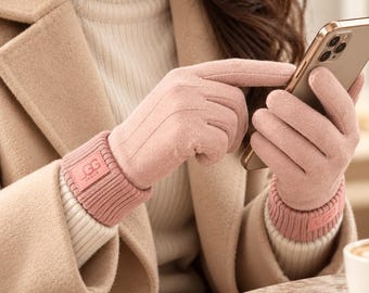 May include: A pair of soft, light pink gloves featuring a ribbed knit cuff with a small rectangular patch displaying the text GG VOGUE. The gloves are worn while interacting with a smartphone, showcasing a smooth, suede-like texture.