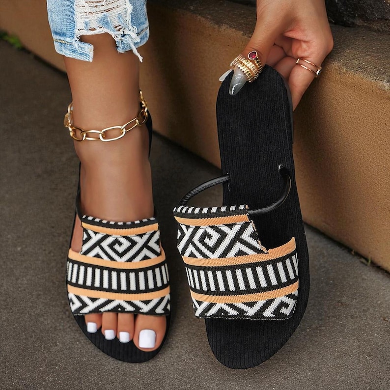 May include: Black sandals with a geometric pattern in orange, white, and black. The sandals have a strap that goes over the top of the foot and a gold chain ankle bracelet. The sandals are open-toed.