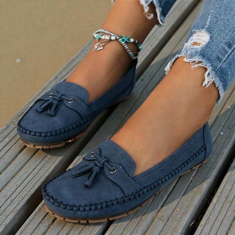 May include: Blue suede loafers with a tassel detail. The shoes have a rounded toe and a stitched border. The soles are brown. The shoes are on a wooden surface.