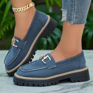 May include: Blue loafers with a chunky black sole and a gold-coloured buckle. The shoes have white stitching and a tan trim around the sole. The shoes are a slip-on style.