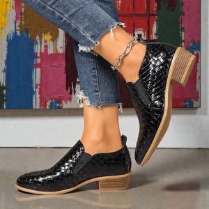 Pu&ograve; includere: Scarpe nere con tacco basso e motivo intrecciato. Le scarpe hanno la punta arrotondata e un piccolo tacco a blocco. I lati delle scarpe hanno pannelli elastici. Le scarpe sono indossate con jeans blu.