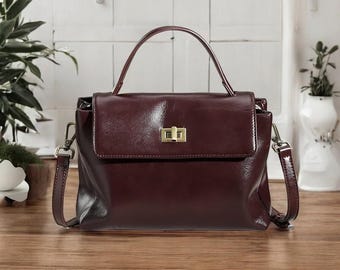 May include: A deep burgundy leather handbag featuring a structured top handle, a fold-over flap with a gold-toned rectangular turn-lock closure, and detachable side straps with metal hardware clips for versatile carrying options.