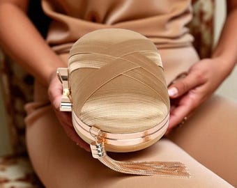 Peut inclure: Une pochette en satin doré avec un motif cousu en croix sur le dessus. La pochette a un cadre en métal doré et une gland suspendu à la fermeture éclair. Le sac est tenu par une personne portant une tenue beige.
