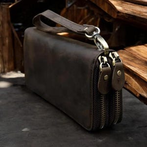 Könnte beinhalten: Eine braune Ledergeldbörse mit drei Reißverschlussfächern und einem Trageriemen. Die Geldbörse hat einen Used-Look und ist aus hochwertigem Leder gefertigt.