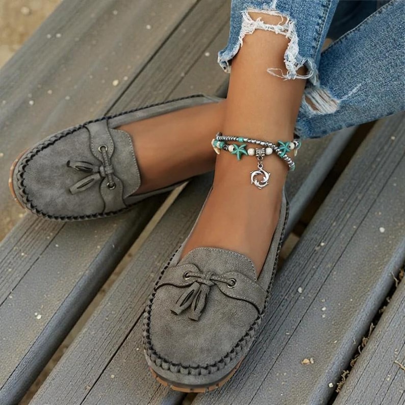 May include: Gray suede loafers with tassel detail. The shoes have a stitched border and a rounded toe. A silver anklet with starfish and a dolphin charm is also visible.